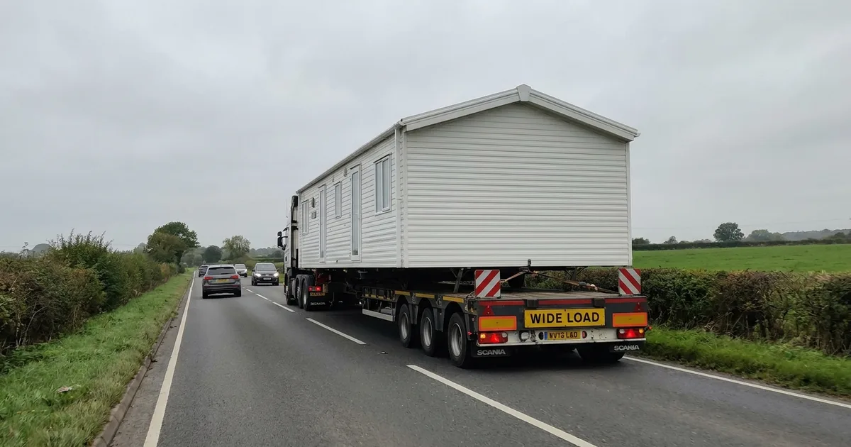 A static caravan strapped onto a specialist low-loader trailer being moved along a UK road